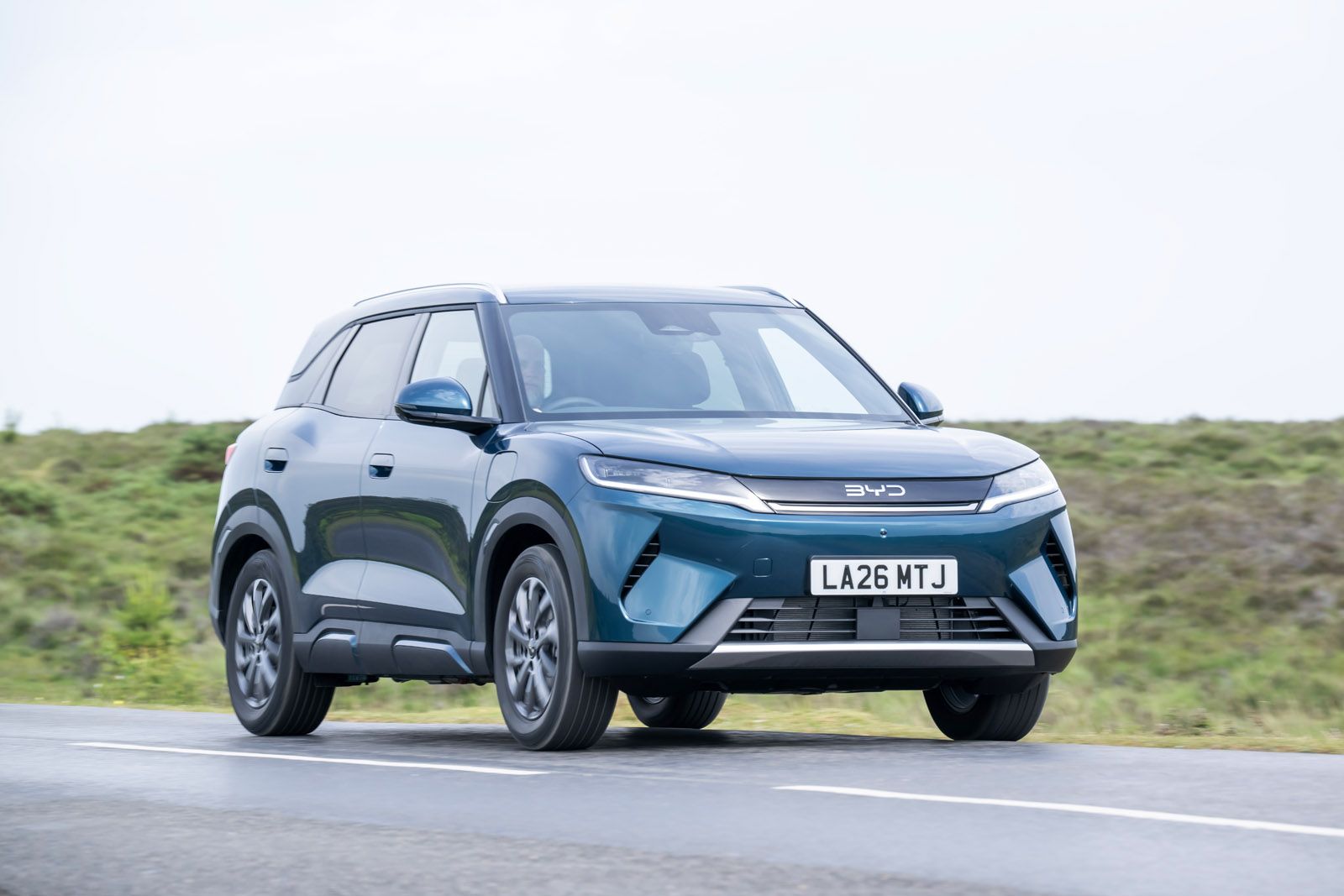 Blue SUV driving on a rural road with grassy landscape, overcast sky. Vehicle has modern design and visible license plate, "LA26 MTJ."