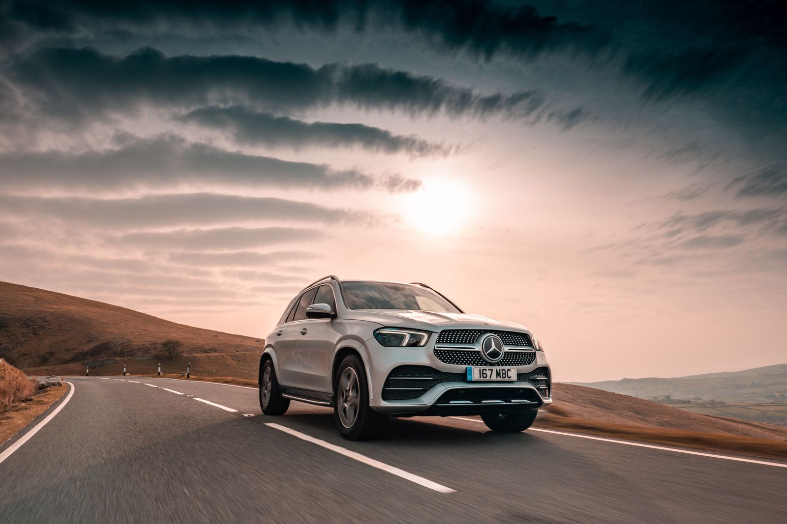 Silver SUV driving on a winding road through a hilly landscape under a dramatic, cloud-filled sky.