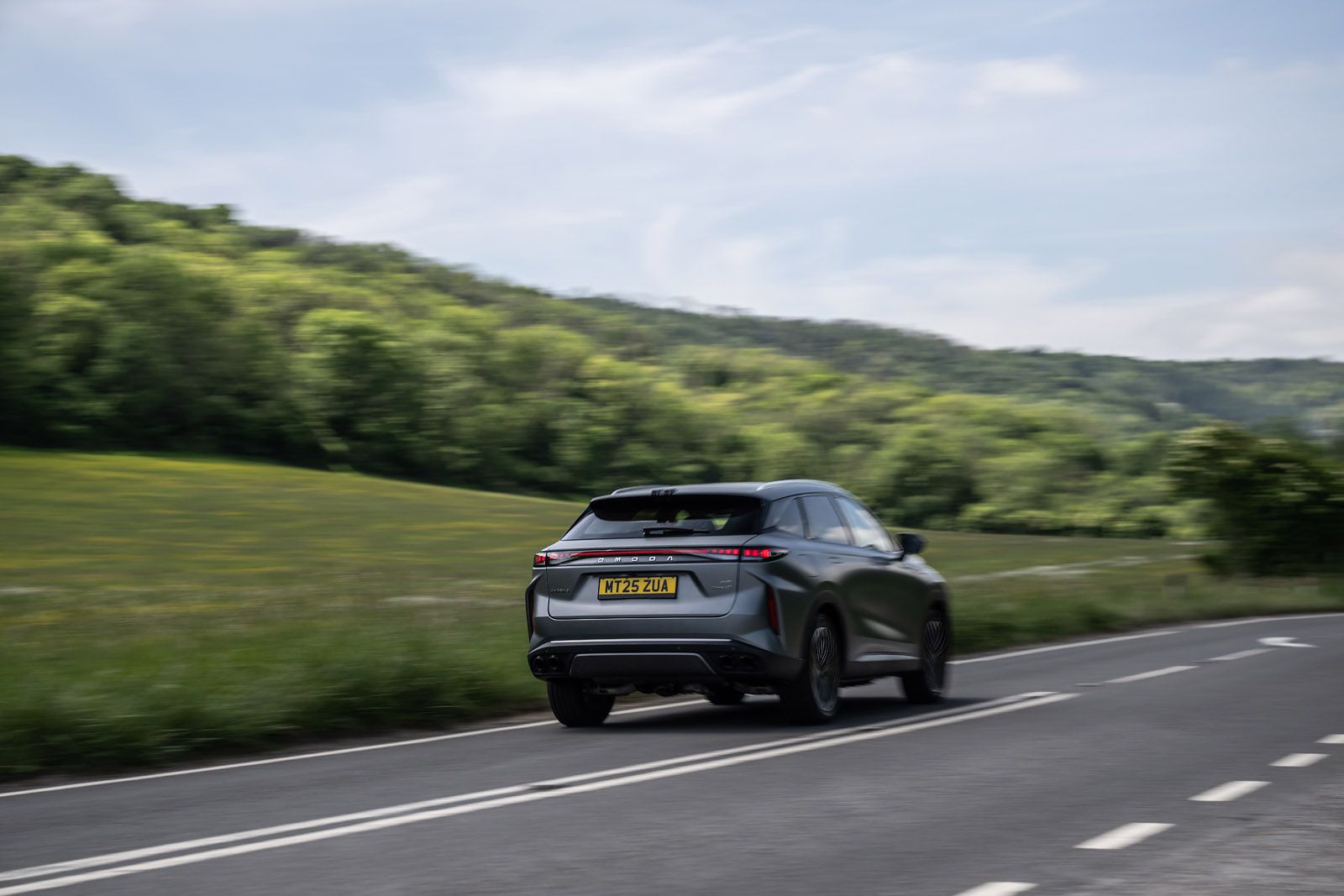 A black SUV drives on a winding road through a lush countryside with hills and greenery in the background under a partly cloudy sky.