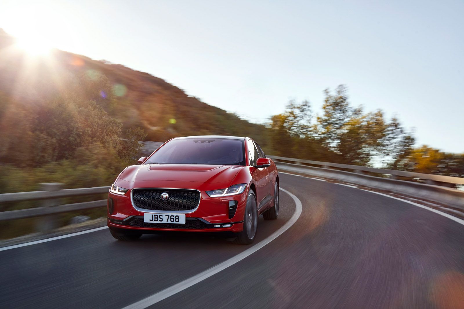 Red car driving on a winding road with sunlight shining from the top left, surrounded by trees and hills.