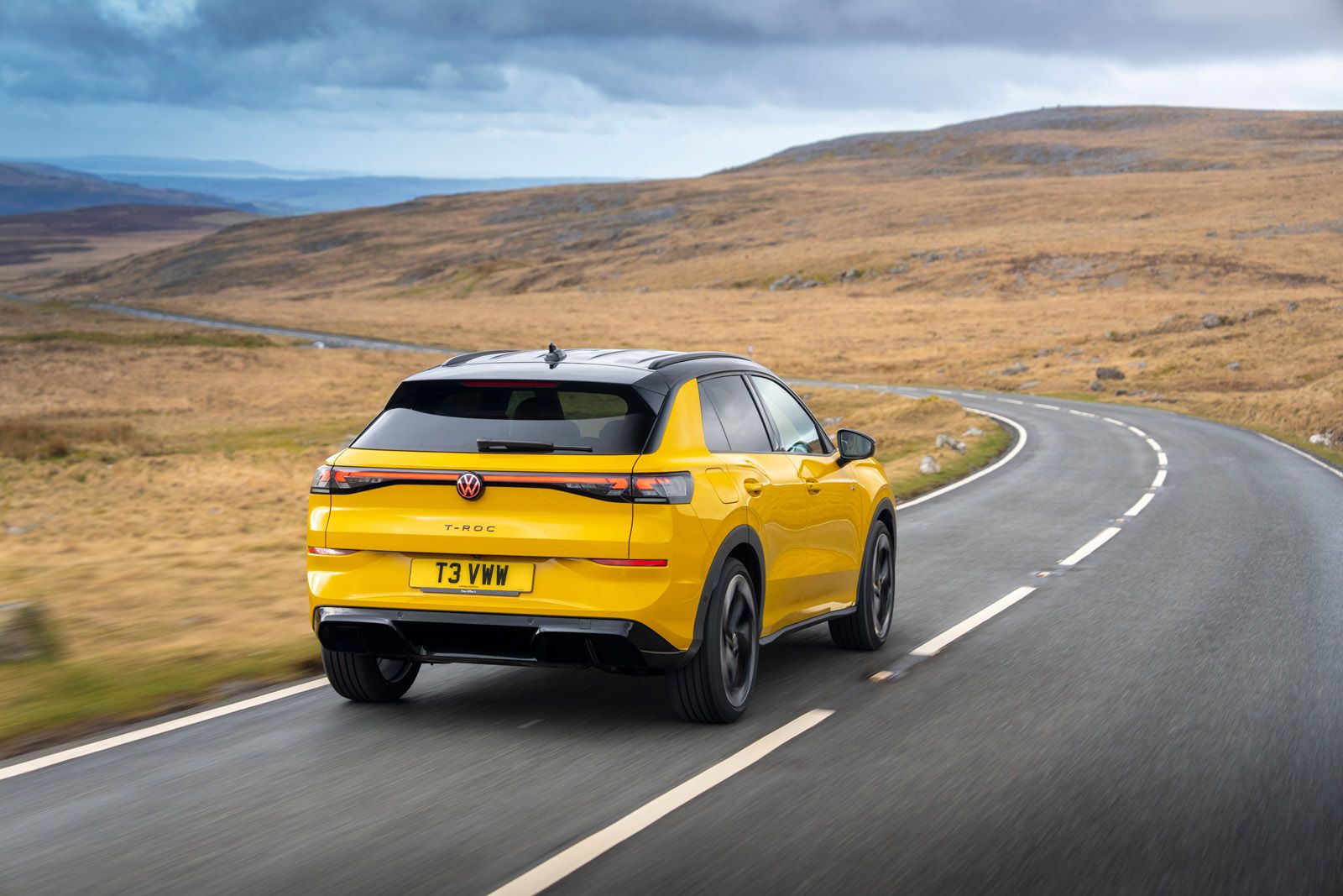Yellow Volkswagen T-Roc on a winding road through a barren landscape, under a cloudy sky.