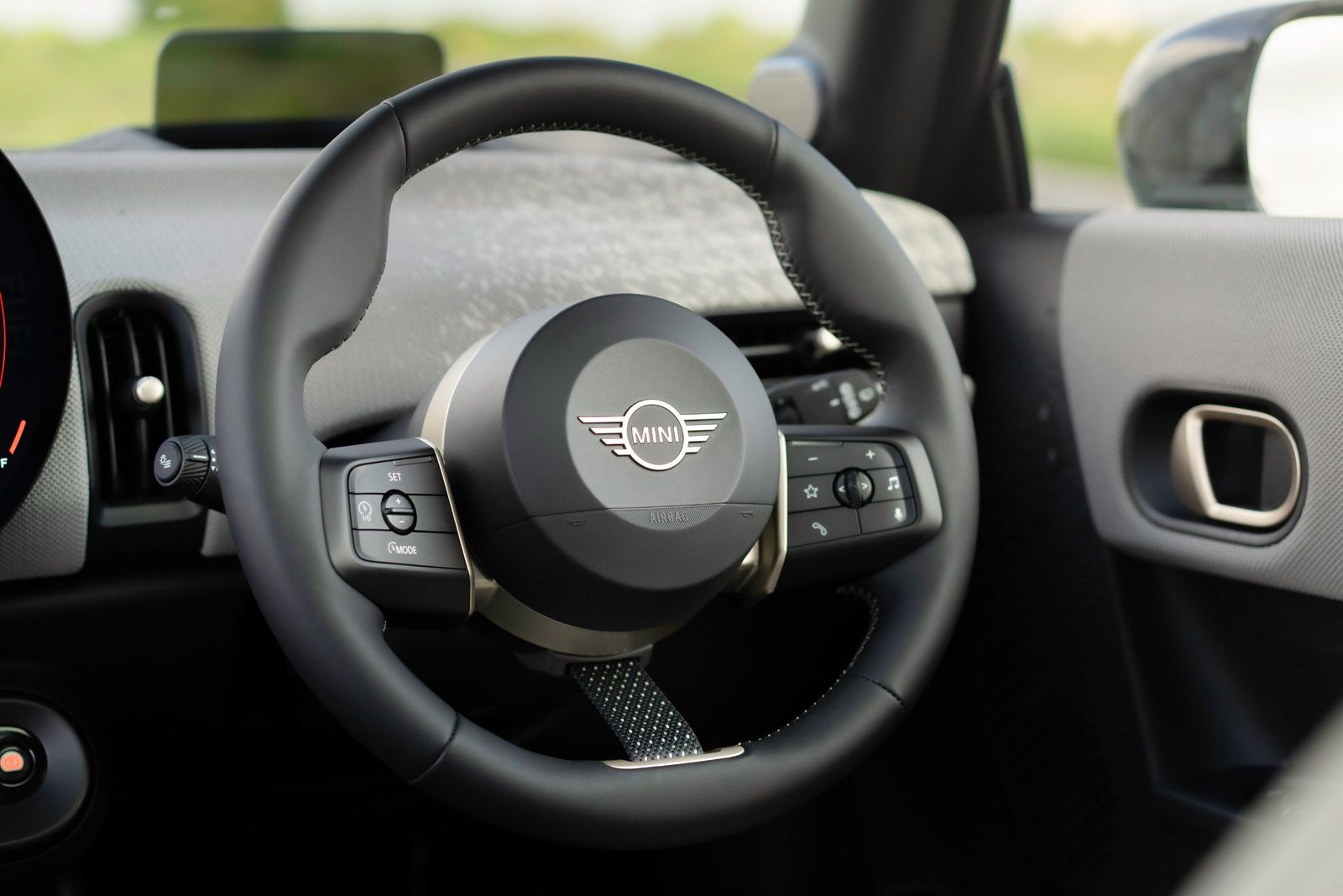 Close-up of a MINI car steering wheel with control buttons, featuring the MINI logo. Black and silver design in a car interior setting.