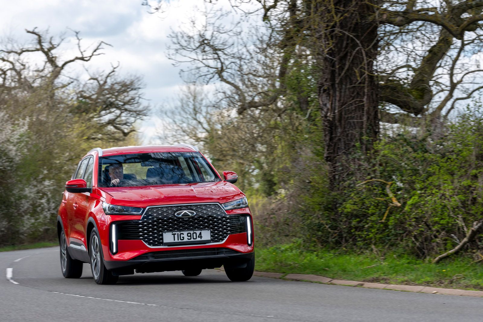 A red SUV driving on a countryside road lined with lush greenery and bare trees under a partly cloudy sky.