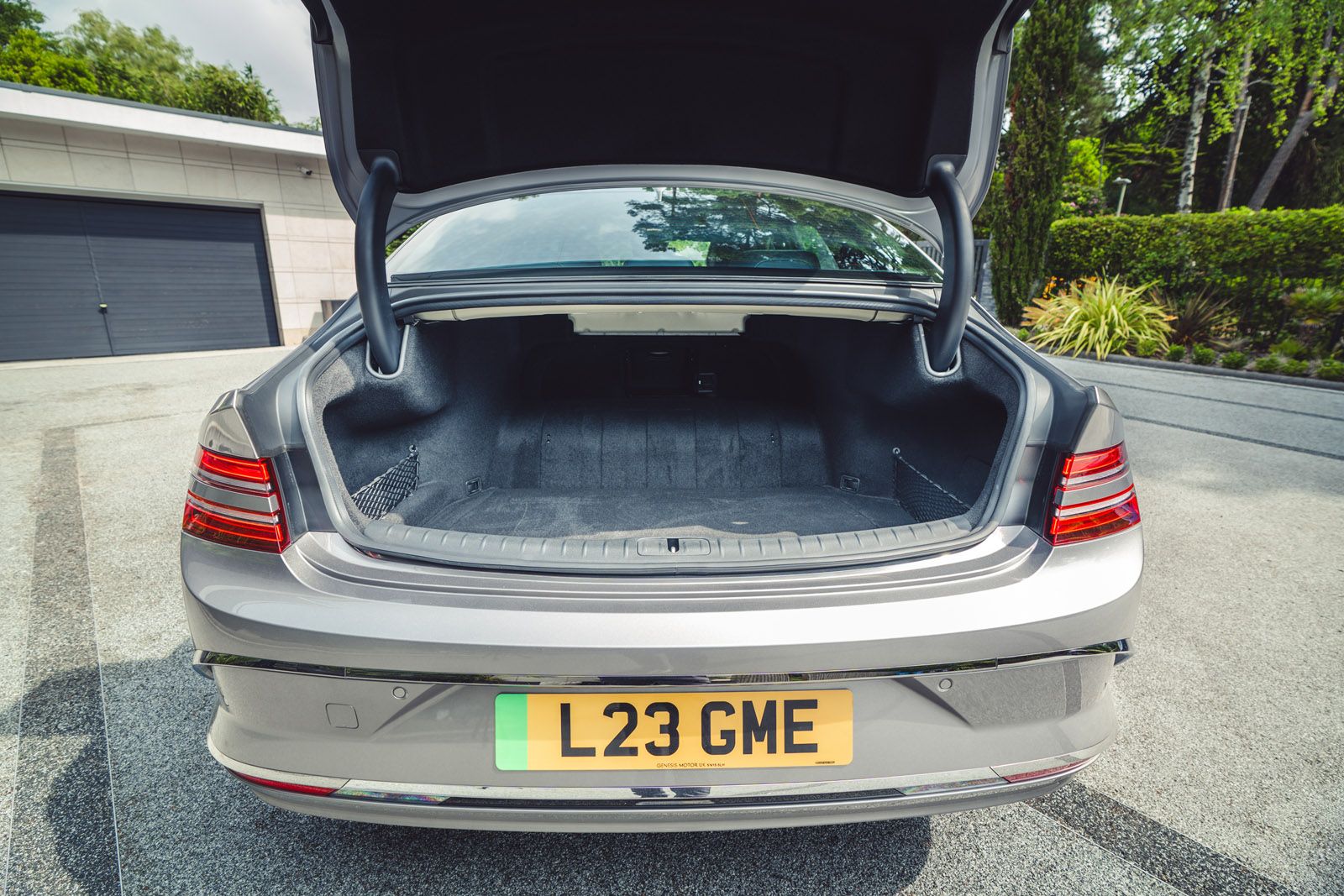 Open trunk of a silver car with license plate L23 GME, parked on a paved driveway in front of a modern house and greenery.