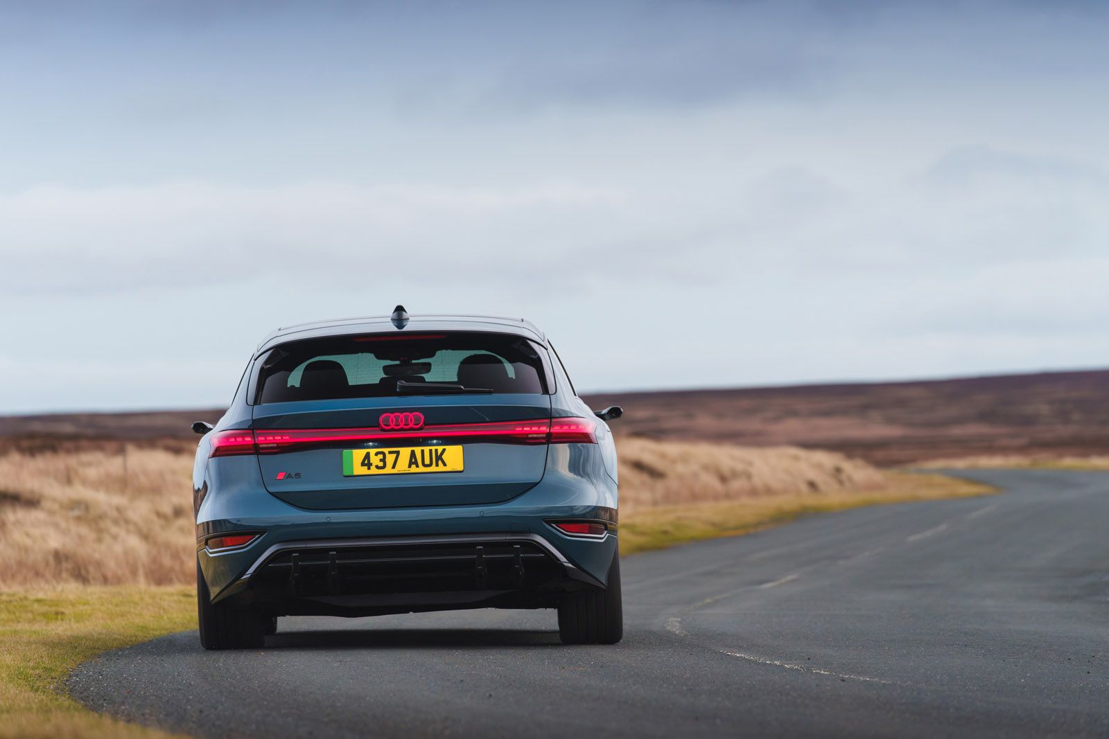 A blue Audi SUV with license plate "437 AUK" drives on an empty rural road under a cloudy sky, surrounded by open fields and moorland.