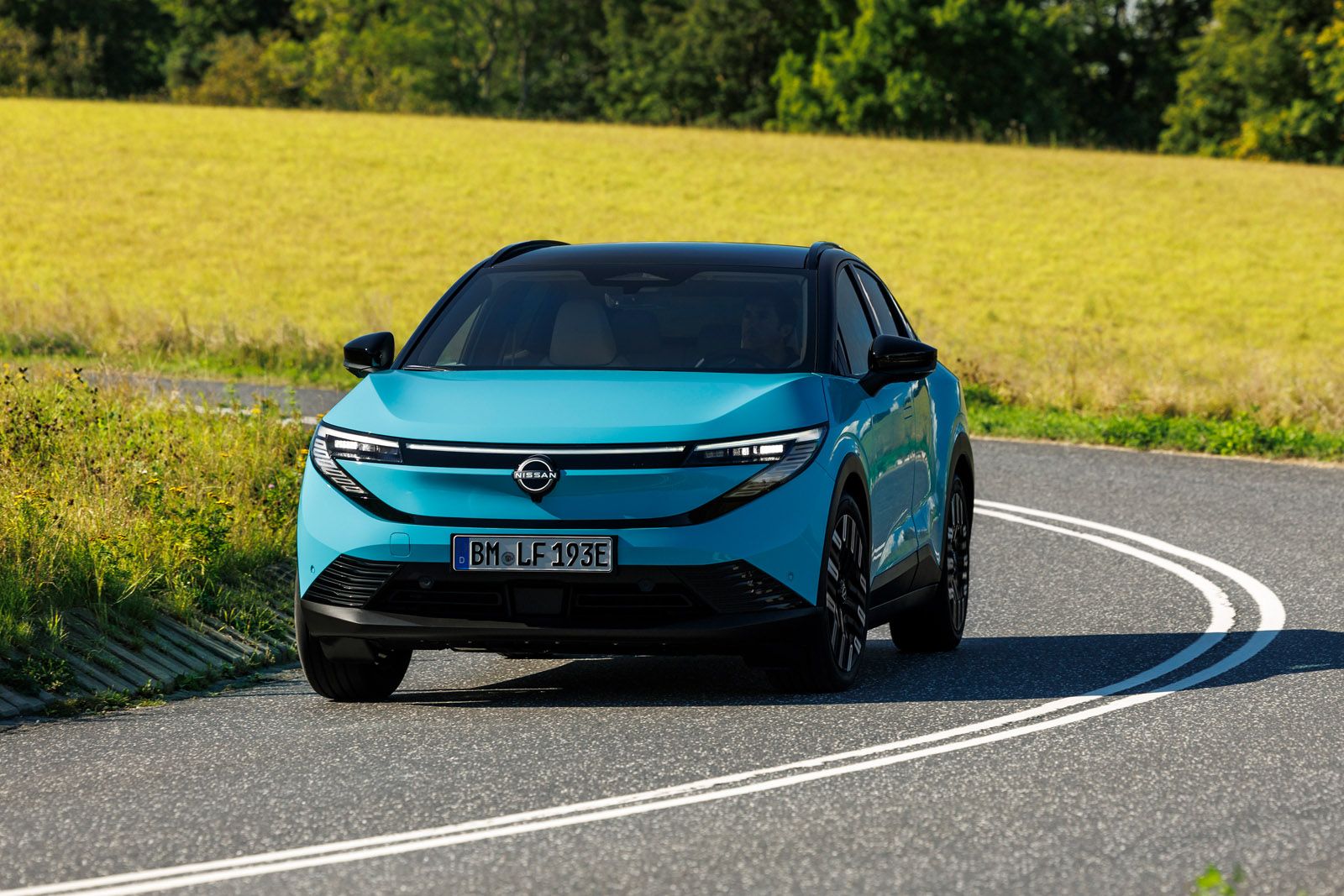 A blue electric Nissan LEAF drives on a curved rural road with lush green fields and trees in the background under a clear sky.