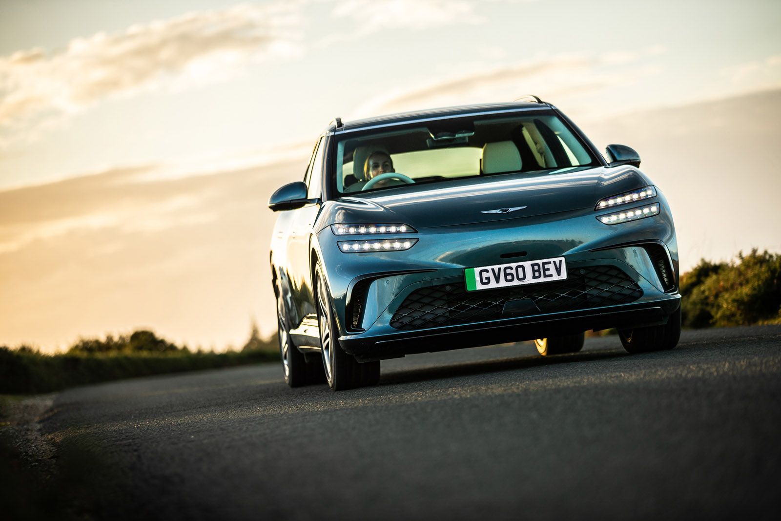 A sleek, blue electric car driving on a scenic road at sunset, with distinctive LED headlights and a "GV60 BEV" license plate.