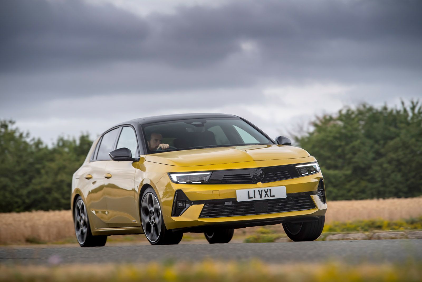 Yellow hatchback car with modern design driving on a country road, overcast sky in the background.