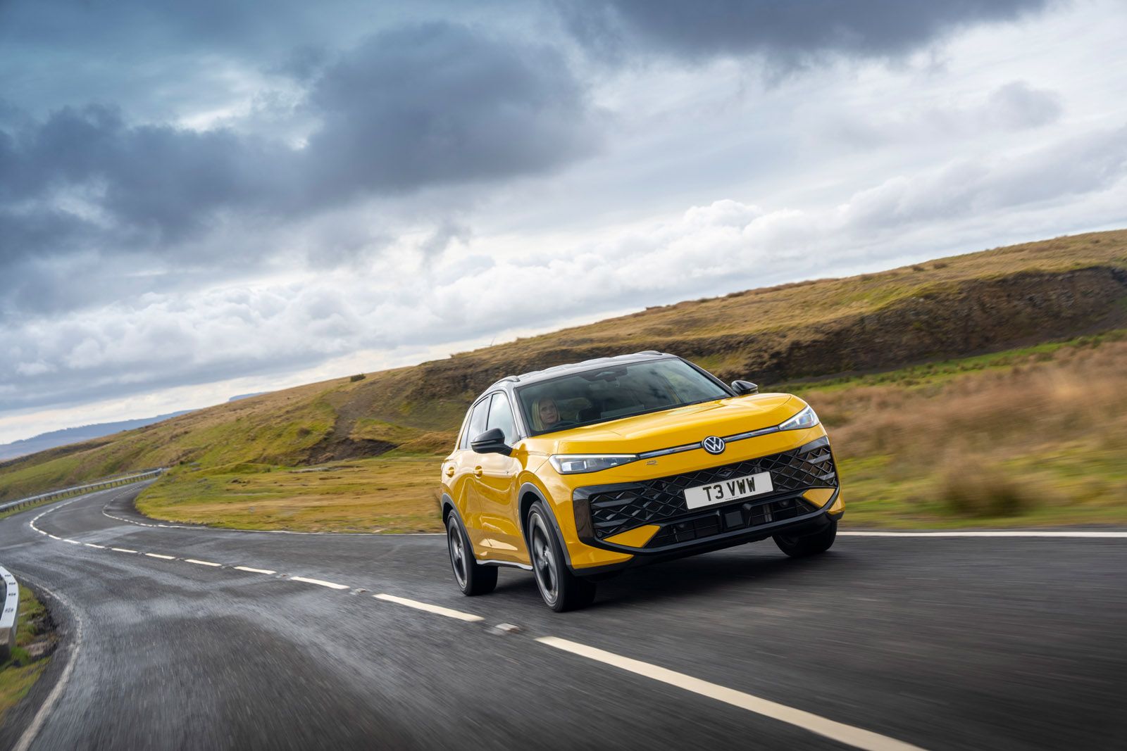 Yellow SUV driving on a curvy countryside road under a cloudy sky, with grass-covered hills in the background.