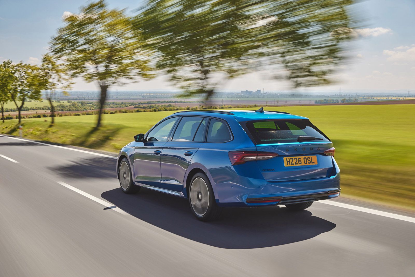 Blue station wagon driving on a scenic rural road, surrounded by green fields and trees, under a clear blue sky.