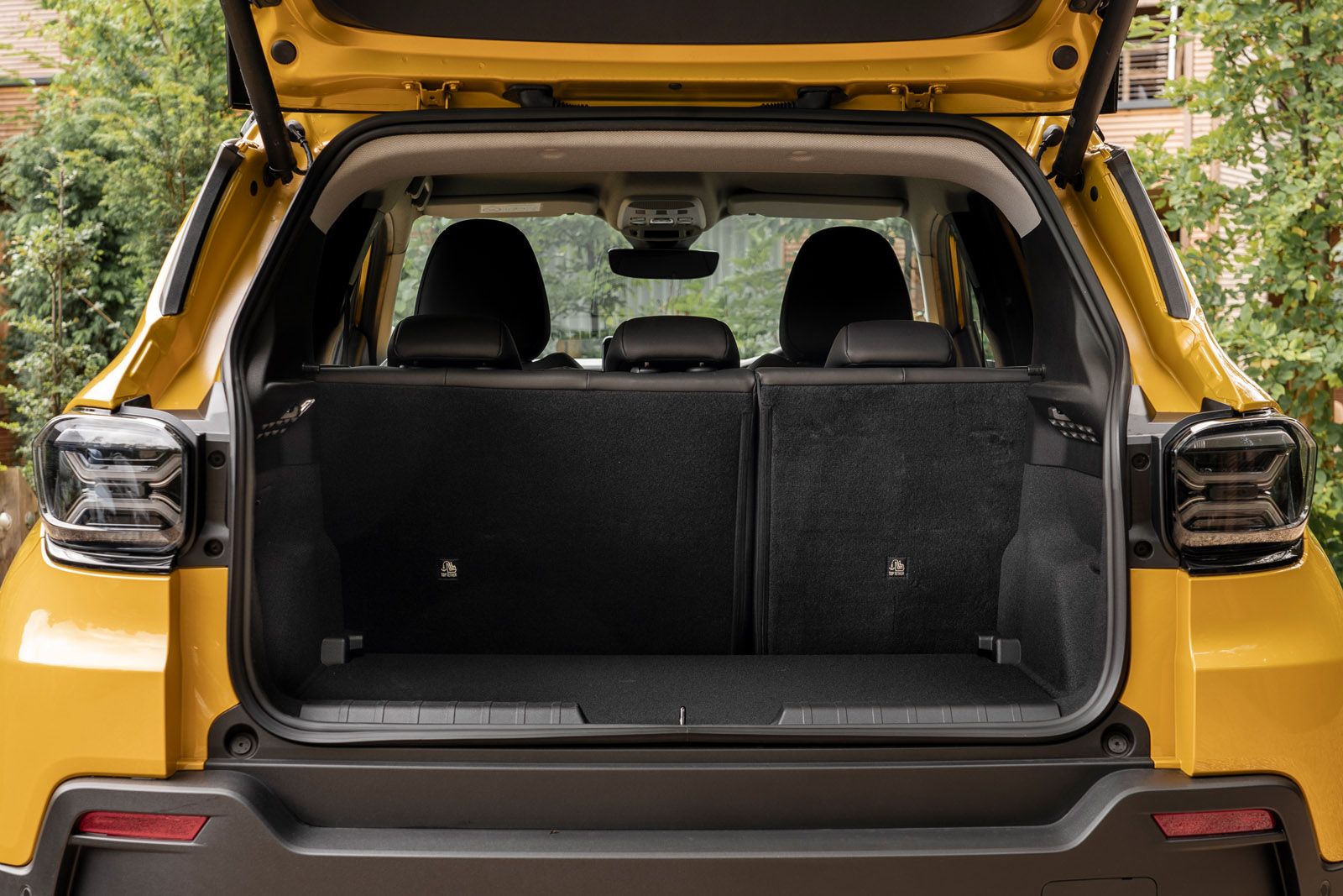 Open trunk of a yellow SUV with rear seats up, revealing a spacious cargo area. Greenery is visible in the background.