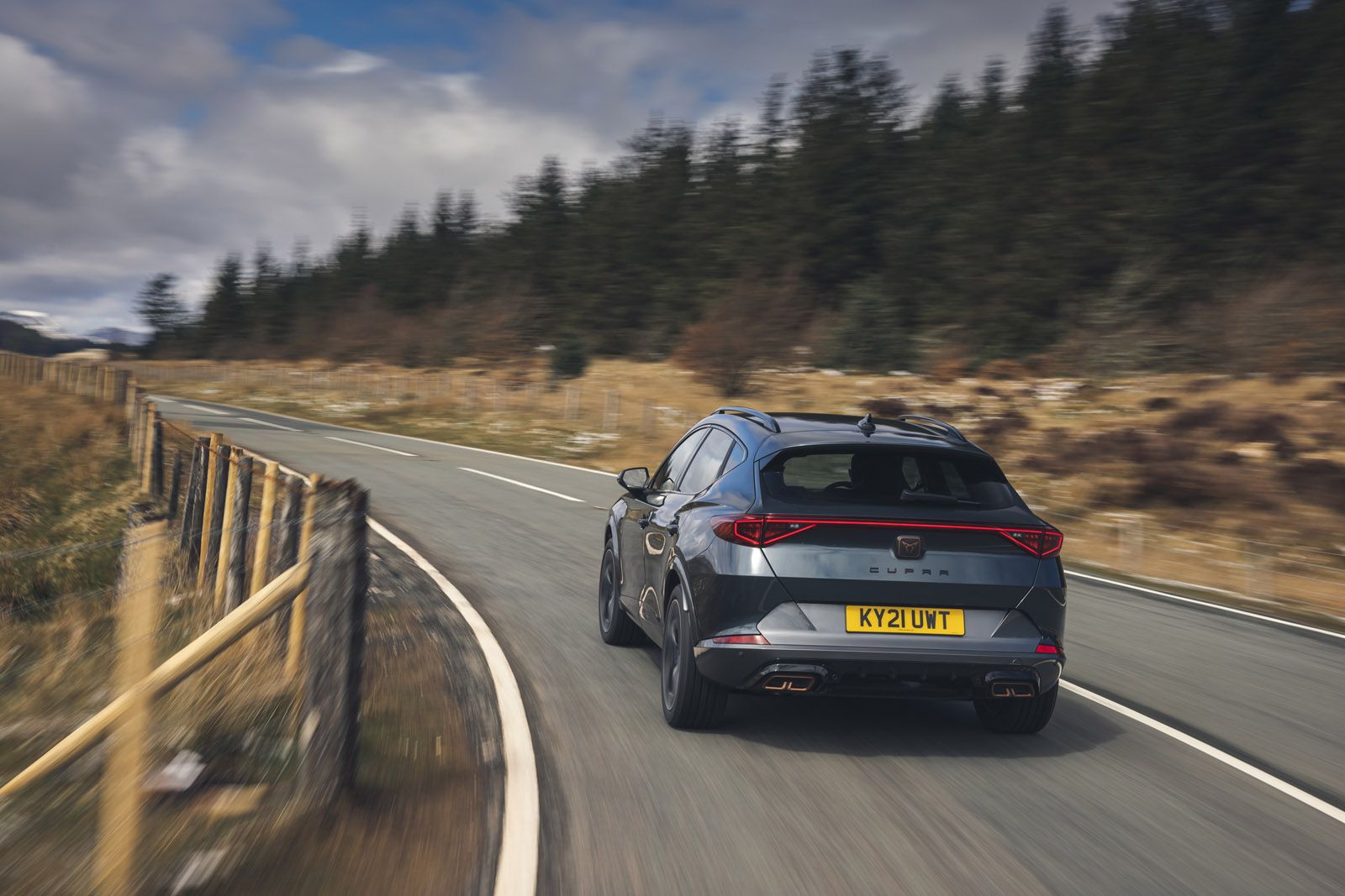 A gray car drives on a winding road through a scenic, hilly landscape with trees, under a cloudy sky.