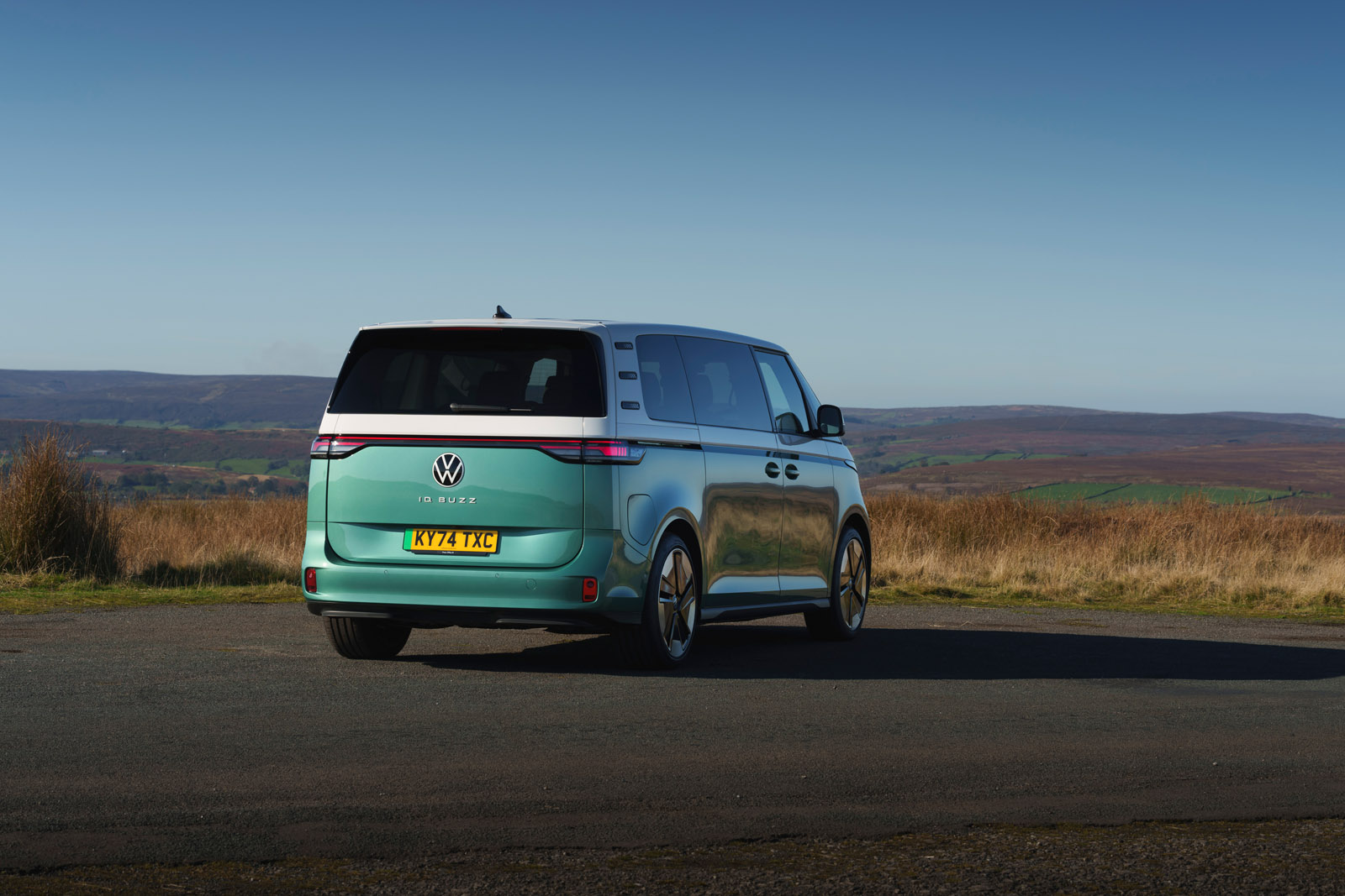 A teal and white Volkswagen van parked on a rural road, with rolling hills and a clear blue sky in the background.