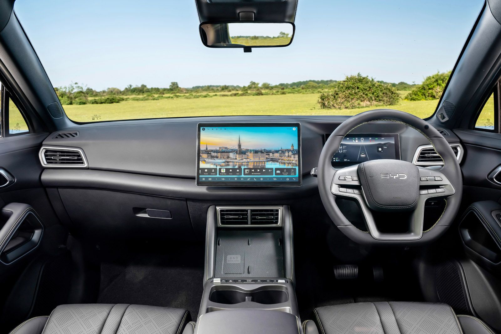 Modern car interior with a touchscreen display, steering wheel, and dashboard controls, overlooking a grassy landscape through the windshield.