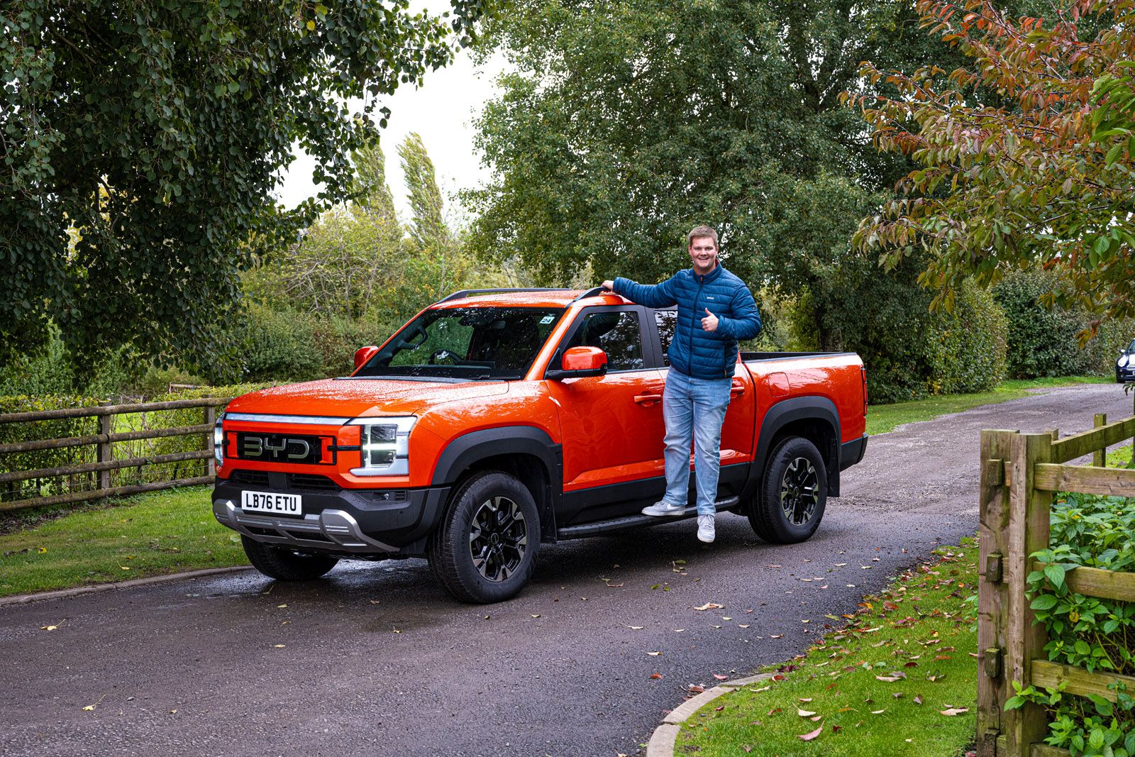 A person in a blue jacket stands beside an orange pickup truck in a lush, leafy countryside setting, giving a thumbs up.
