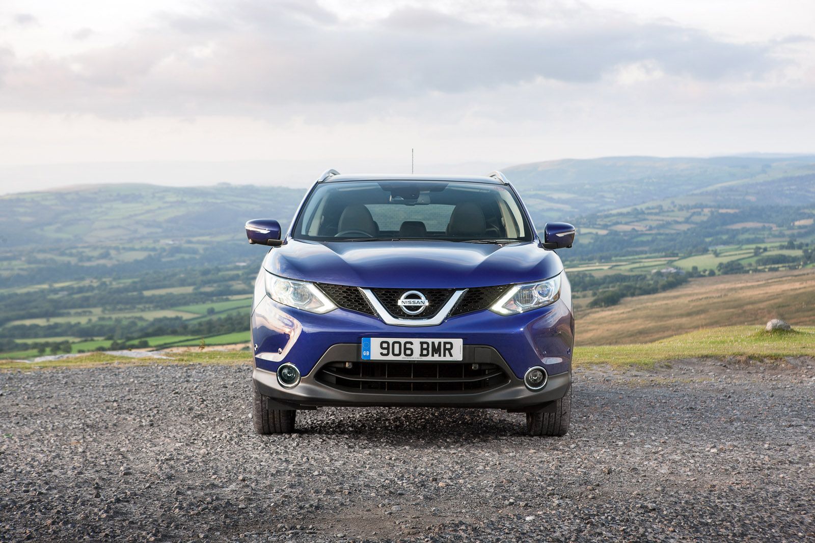A blue Nissan car parked on a gravel road overlooking a scenic, green landscape with rolling hills under a cloudy sky.