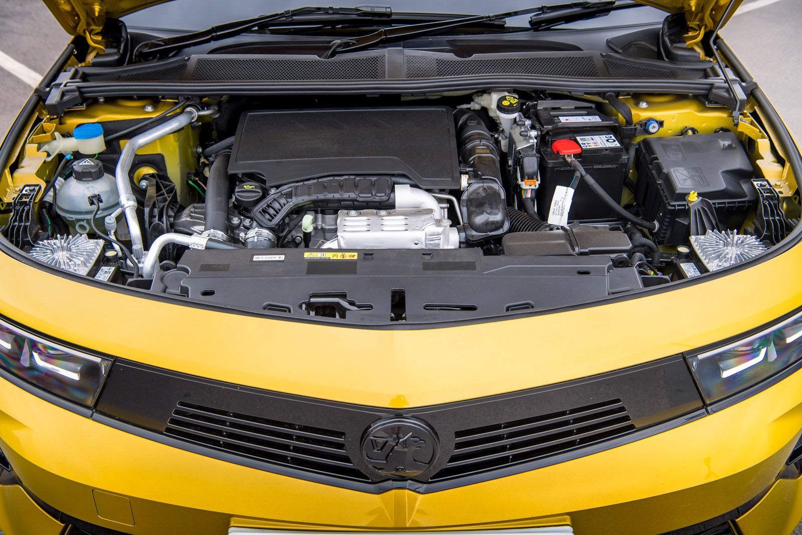 The image shows the engine compartment of a yellow car with various components, including the battery and fluid reservoirs, clearly visible.