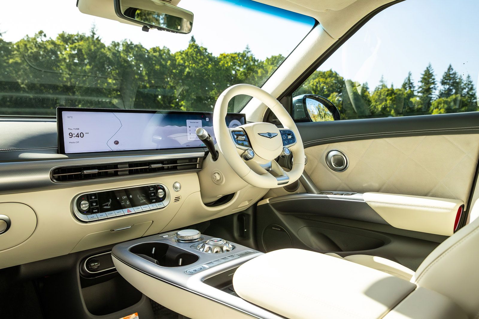 Modern car interior with beige leather seats, sleek dashboard, digital displays, and a wooded landscape visible through the windows.