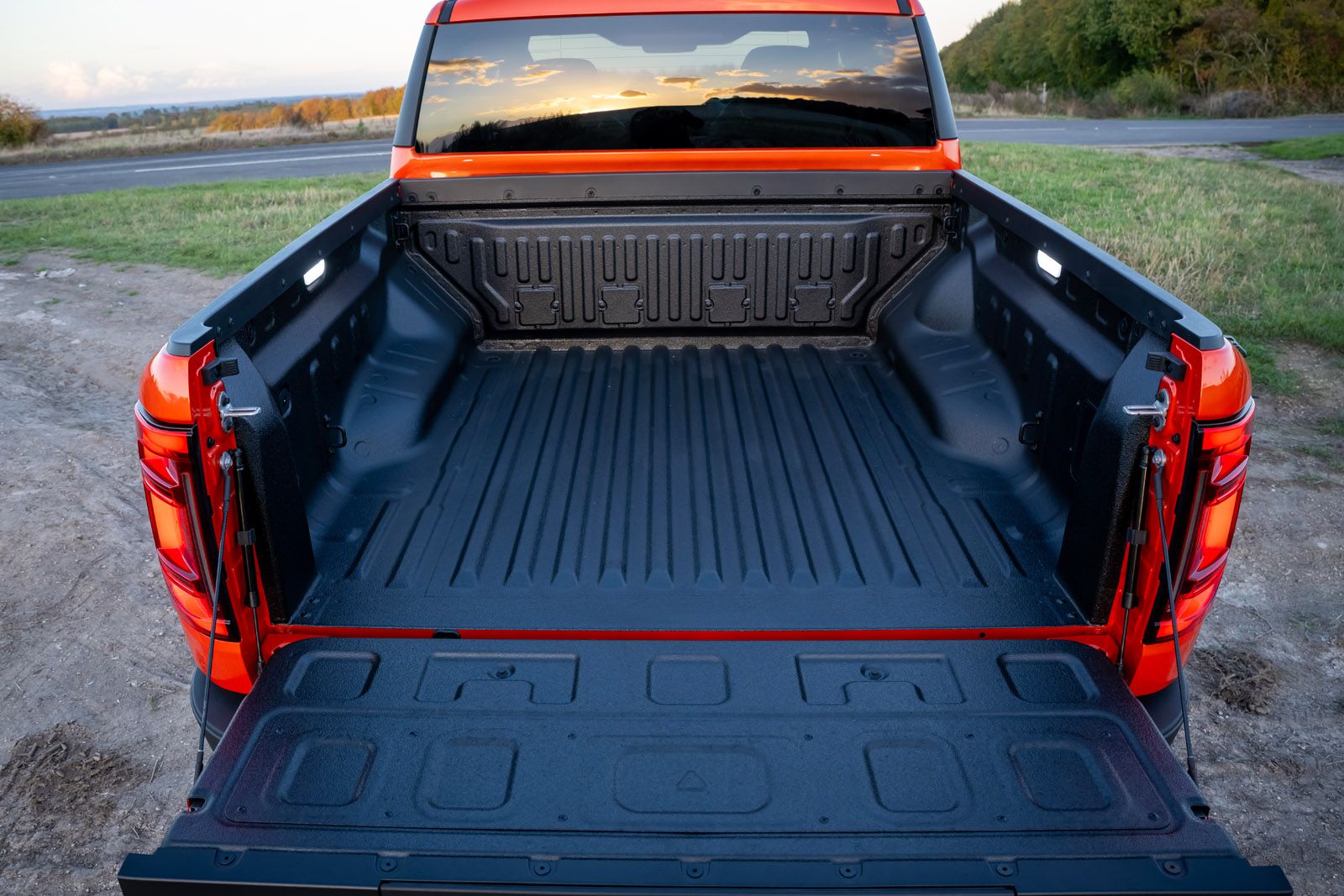The open bed of an orange pickup truck with a black liner, parked outdoors near a road with green fields and trees in the background.