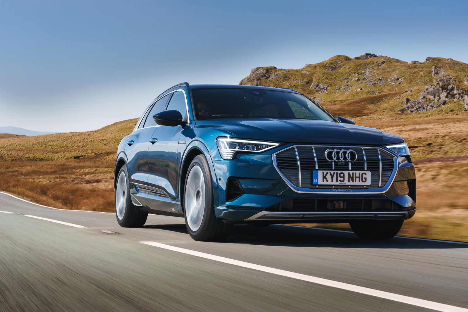 A blue Audi e-tron SUV driving on a scenic road with grassy hills and a clear sky in the background.