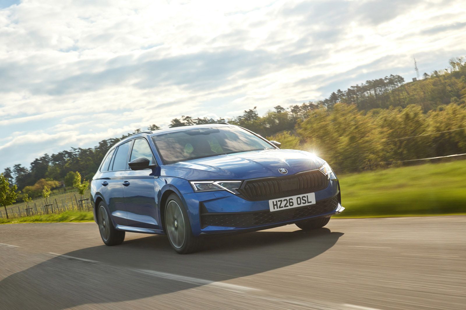 Blue car driving on a rural road with trees in the background and a bright sky above.