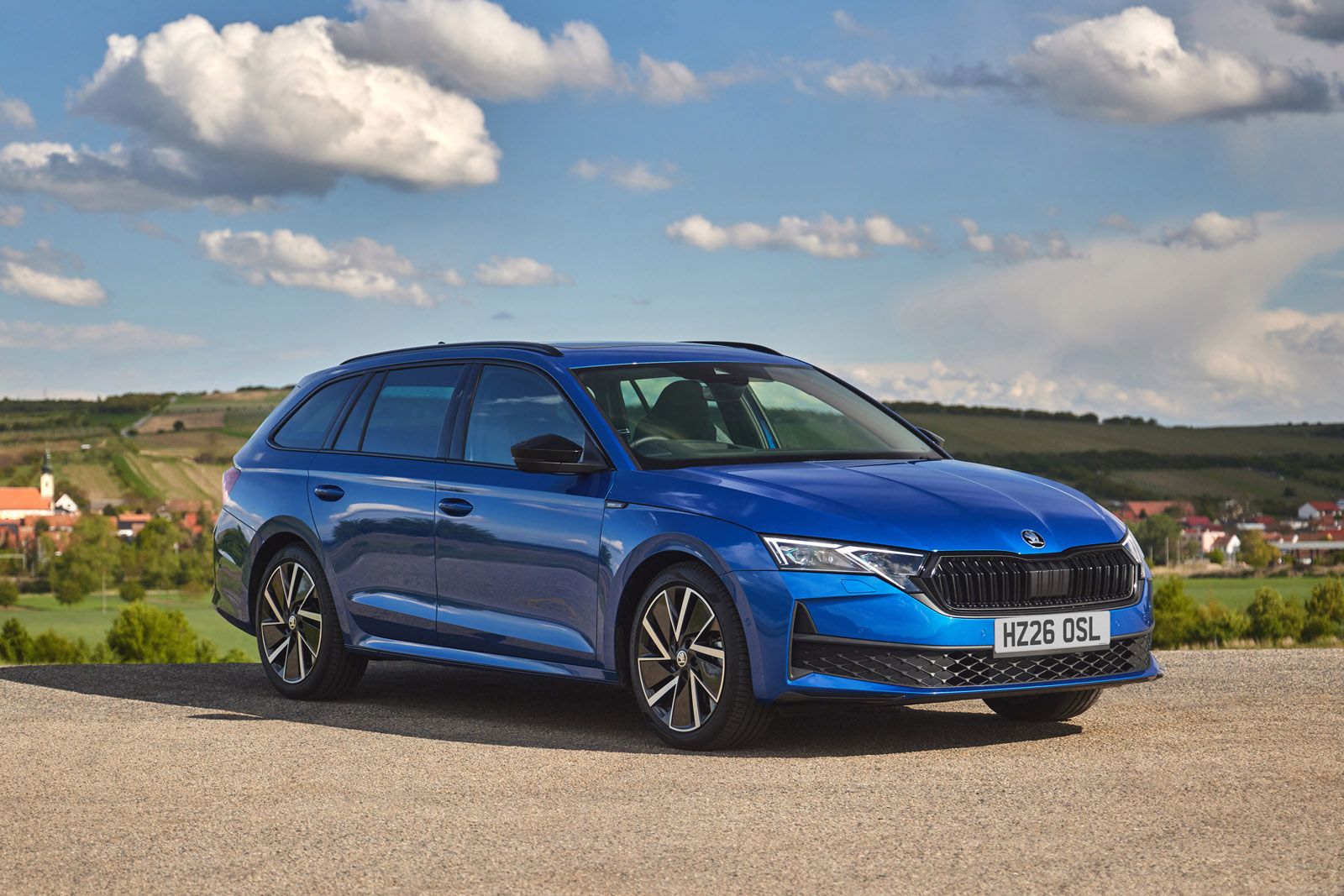 Blue station wagon parked in a scenic rural area, under a partly cloudy sky with fields and houses in the background.