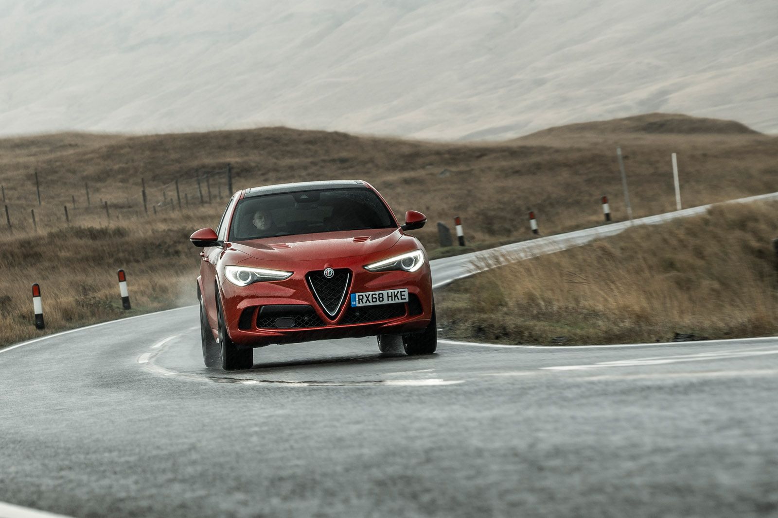 Red sports car driving on a winding, wet road through a barren, hilly landscape under an overcast sky.