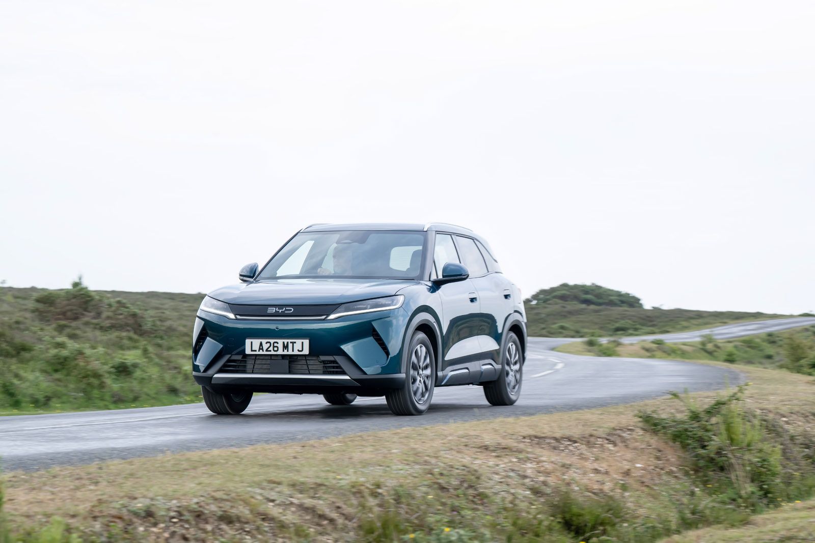 A blue SUV drives on a winding countryside road under an overcast sky.