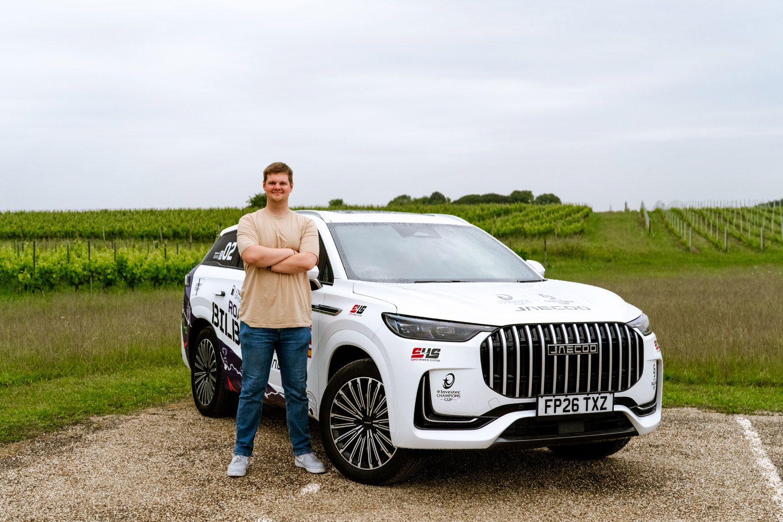Man standing with arms crossed next to a white SUV with decals, set against a vineyard backdrop on a cloudy day.