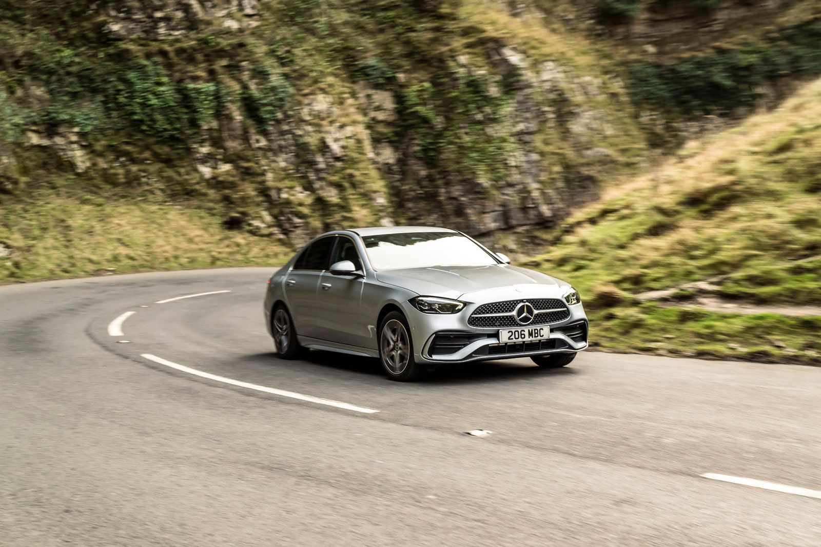 Silver Mercedes-Benz car driving on a winding mountain road with grassy hills and rocky cliffs in the background.
