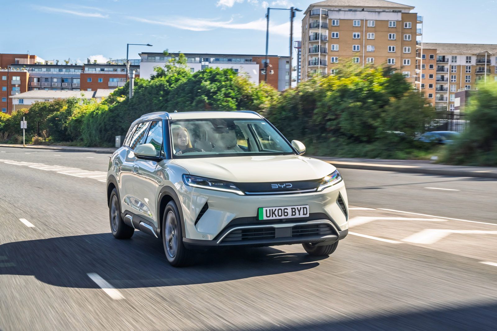 A silver SUV driving on a city road with blurred motion, surrounded by greenery and buildings under a partly cloudy sky.