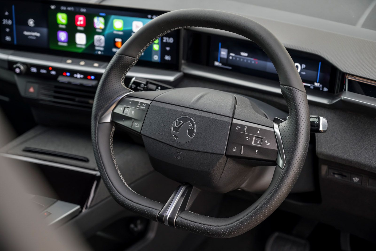 Close-up of a car's steering wheel with control buttons, featuring a modern digital dashboard displaying apps and control settings in the background.