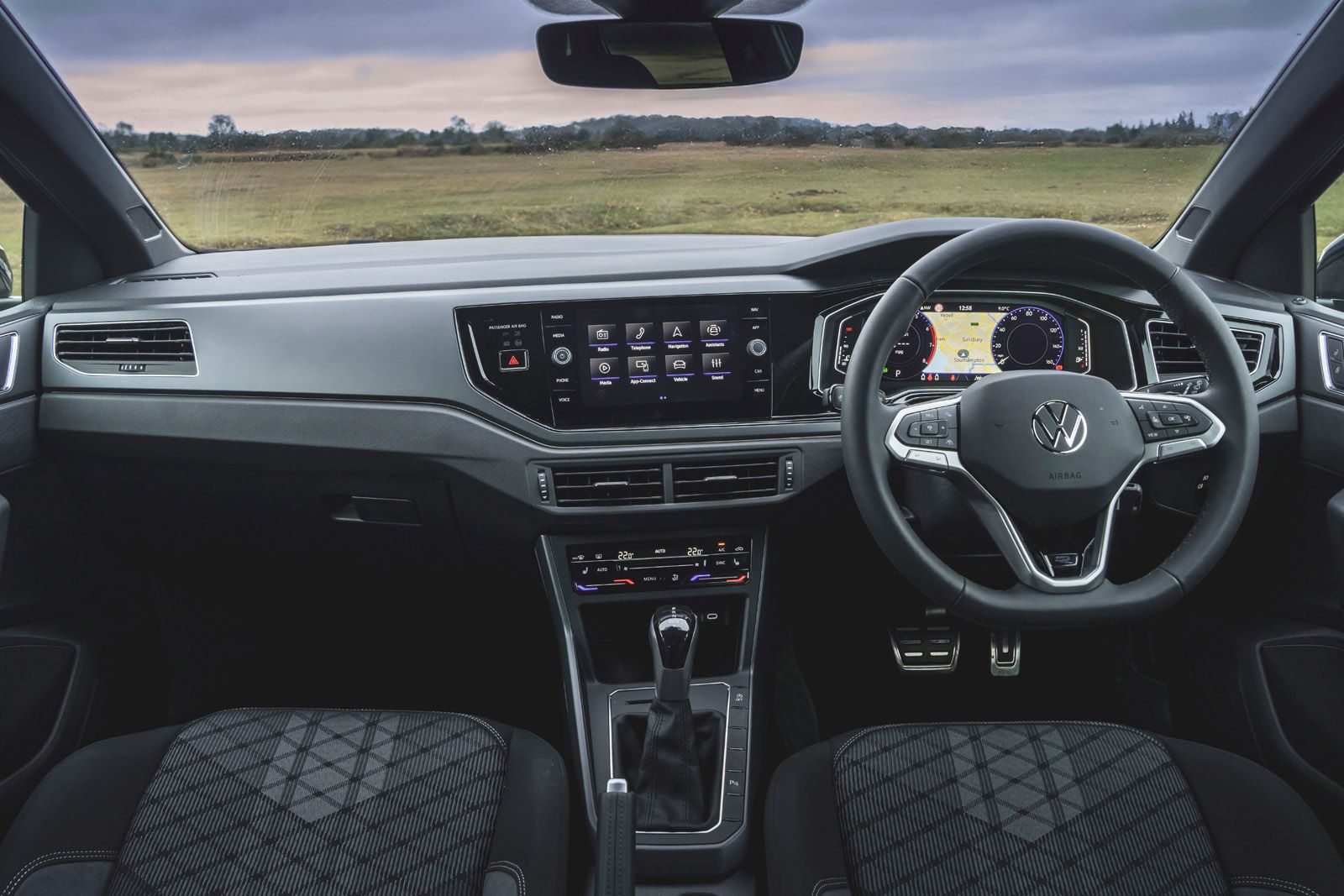 Interior of a Volkswagen car showing the dashboard with central touchscreen, digital display, and steering wheel, set against a grassy landscape.