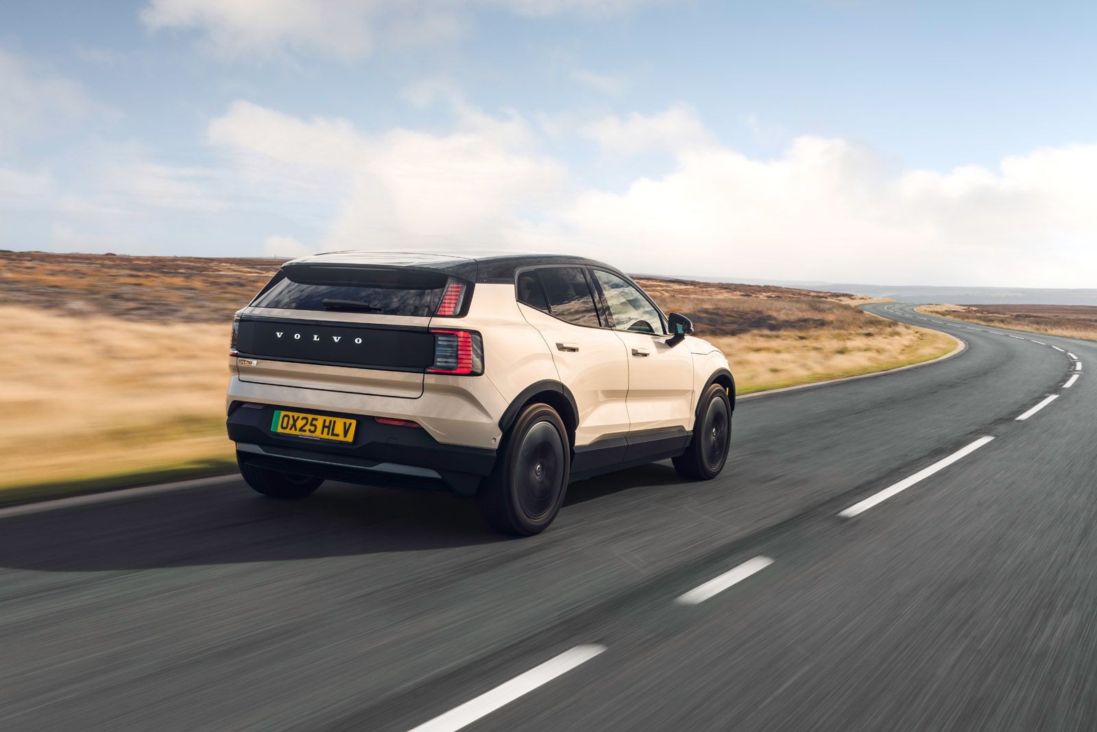 A beige Volvo SUV driving on a winding rural road with grassy fields under a partly cloudy sky.
