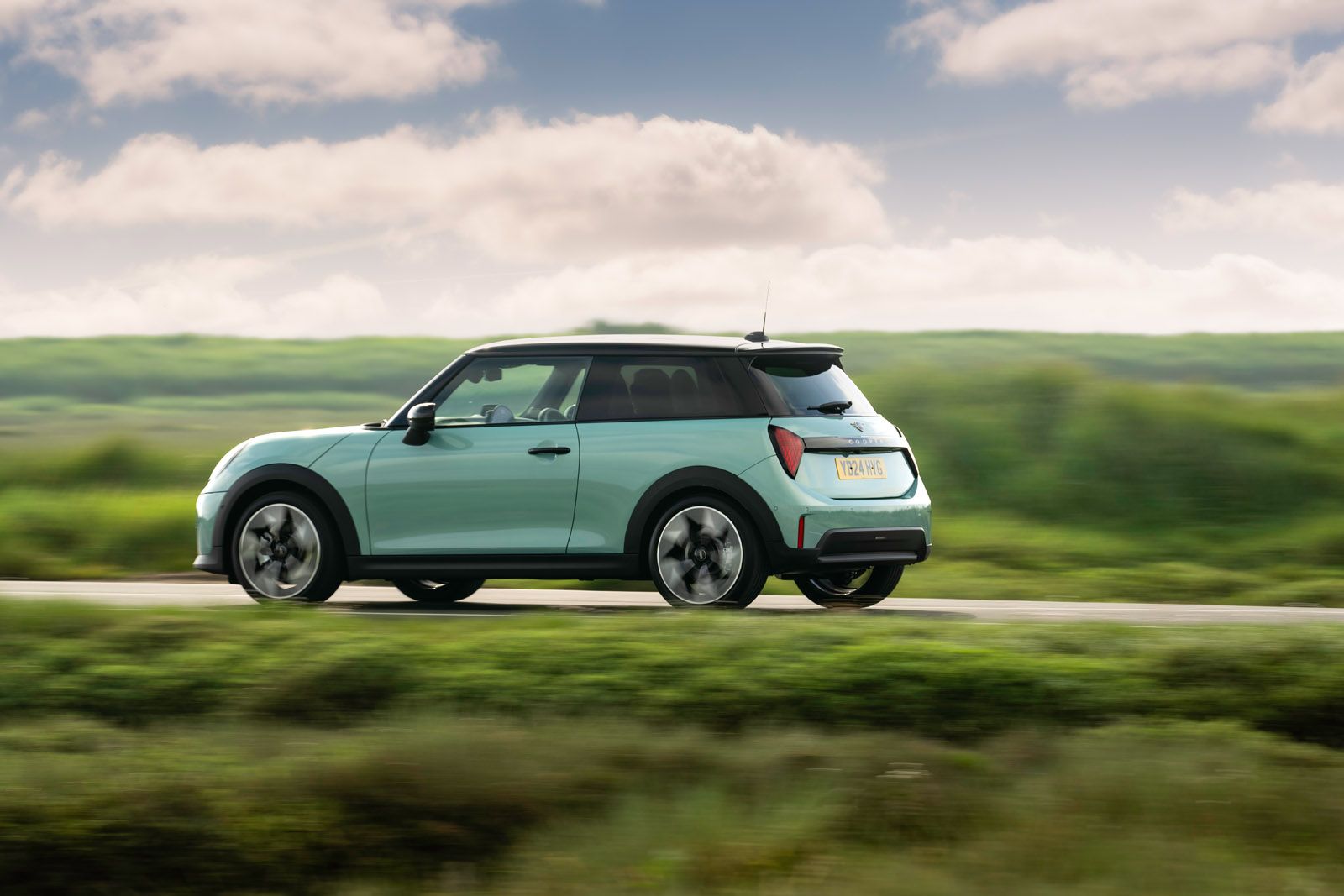 A turquoise compact car driving on a road with blurred green fields and a cloudy blue sky in the background.