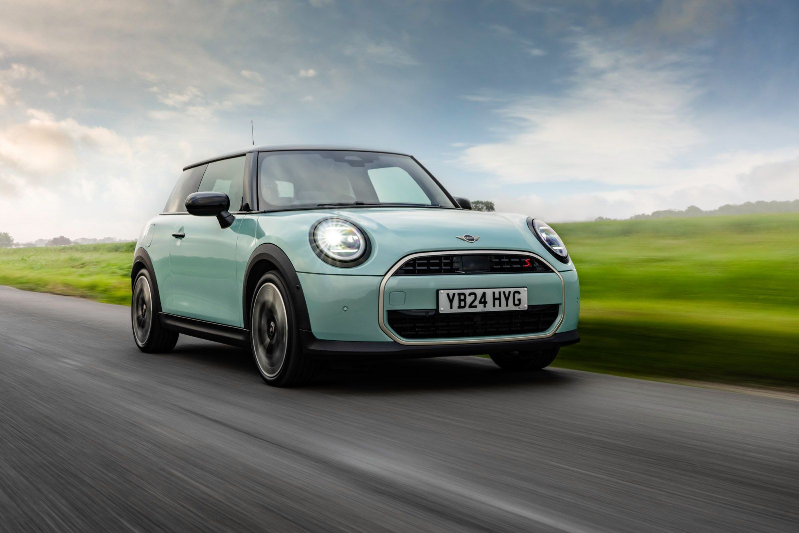 Light blue Mini Cooper driving on a countryside road with green fields and a cloudy sky in the background.