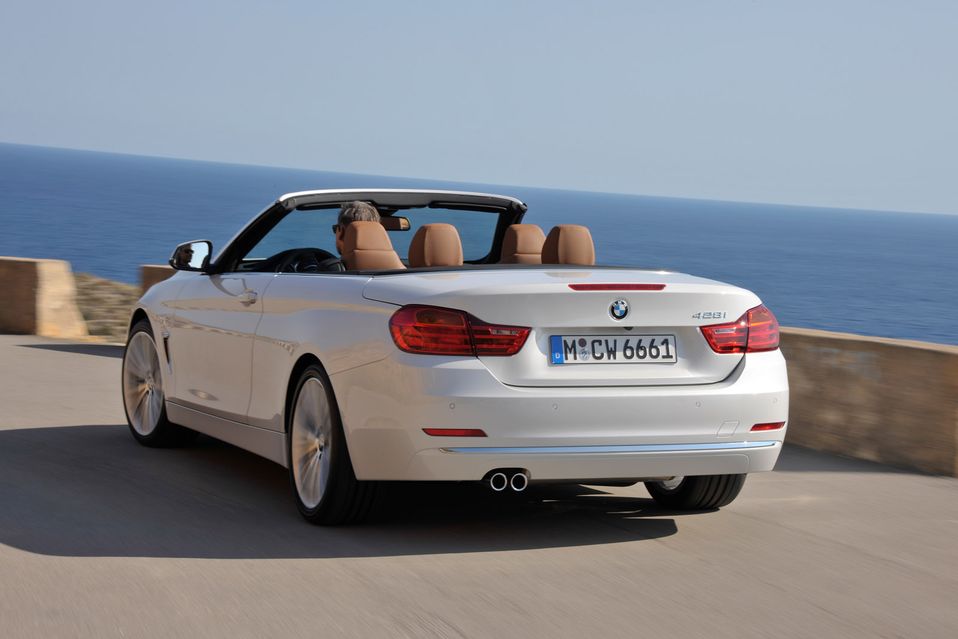 White convertible driving on a coastal road, top down, with a view of the sea and clear sky in the background.