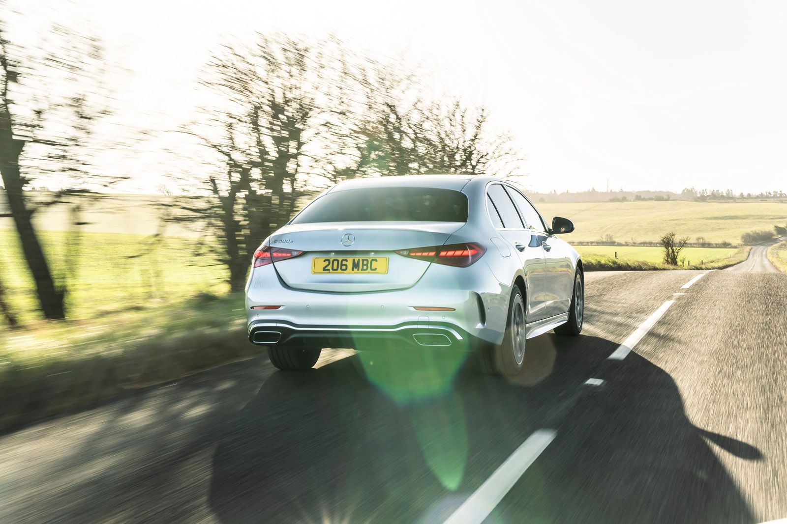 Silver car with license plate "206 MBC" drives on a sunny rural road, surrounded by fields and leafless trees.