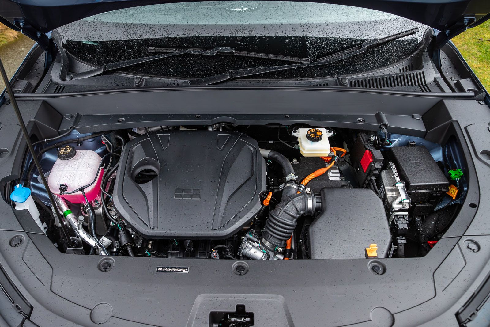 Car engine bay under an open hood, showing various components with some rain droplets on surfaces, indicating wet weather.