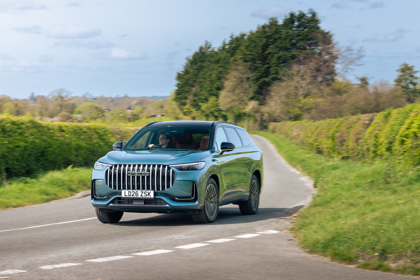 A green SUV driving on a rural road surrounded by lush greenery and trees under a partly cloudy sky.