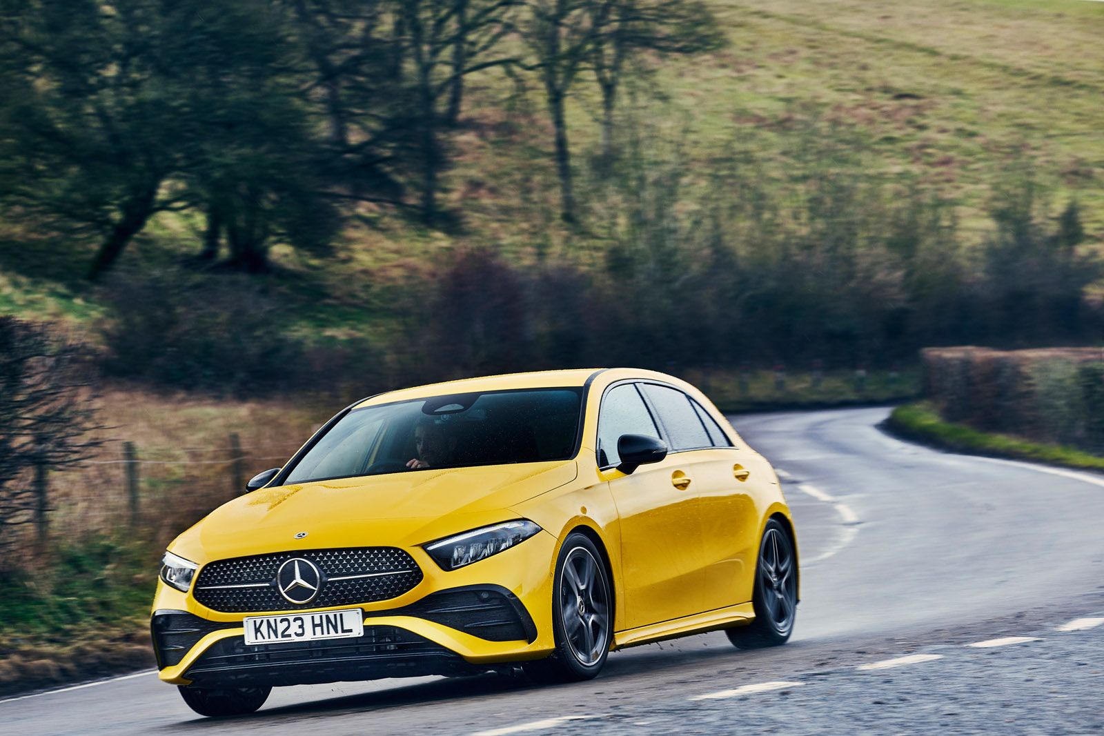 Yellow Mercedes-Benz car driving on a curvy road through a countryside landscape with grassy hills and leafless trees.