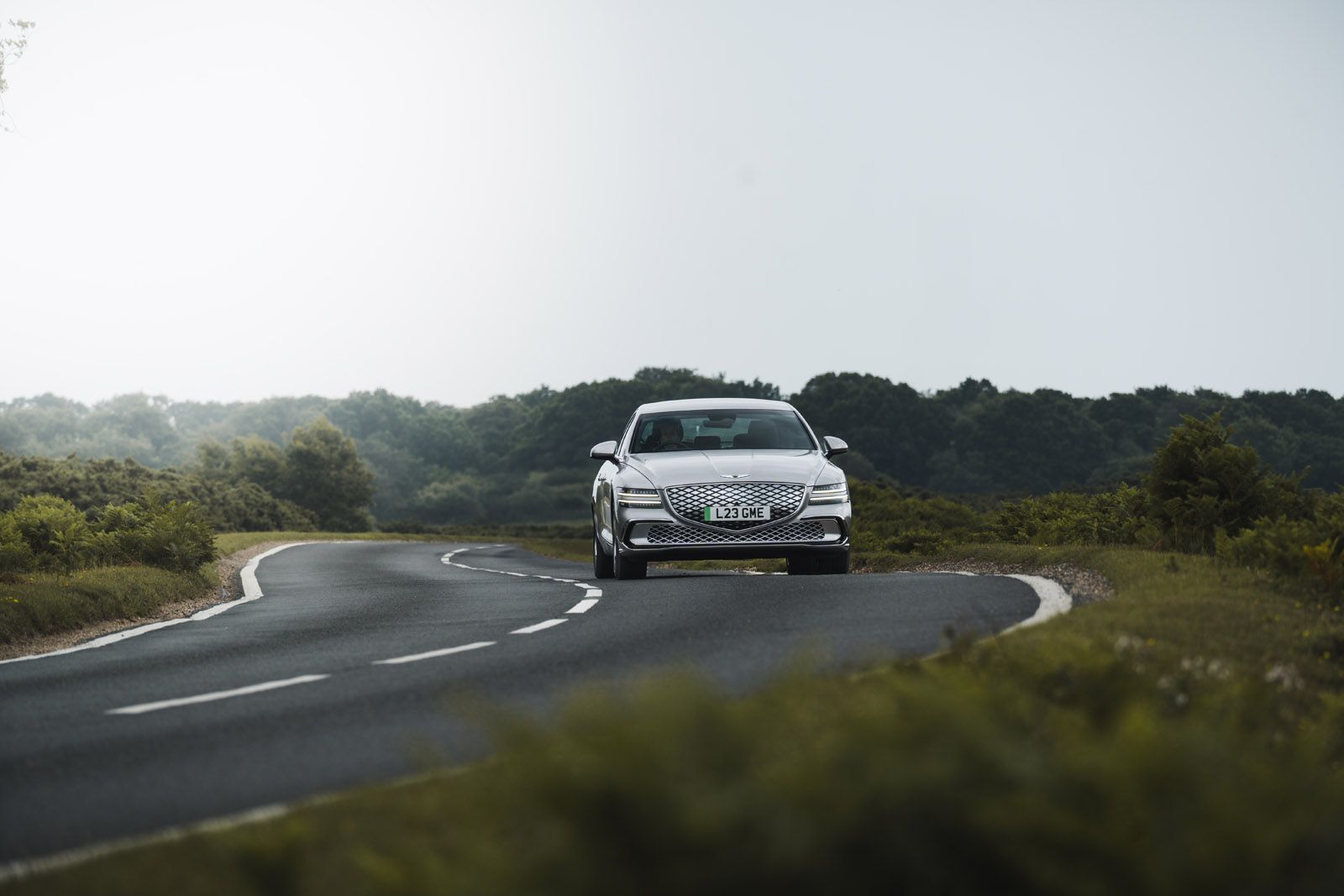 Silver sedan driving on a winding, empty road through a lush green landscape under an overcast sky.