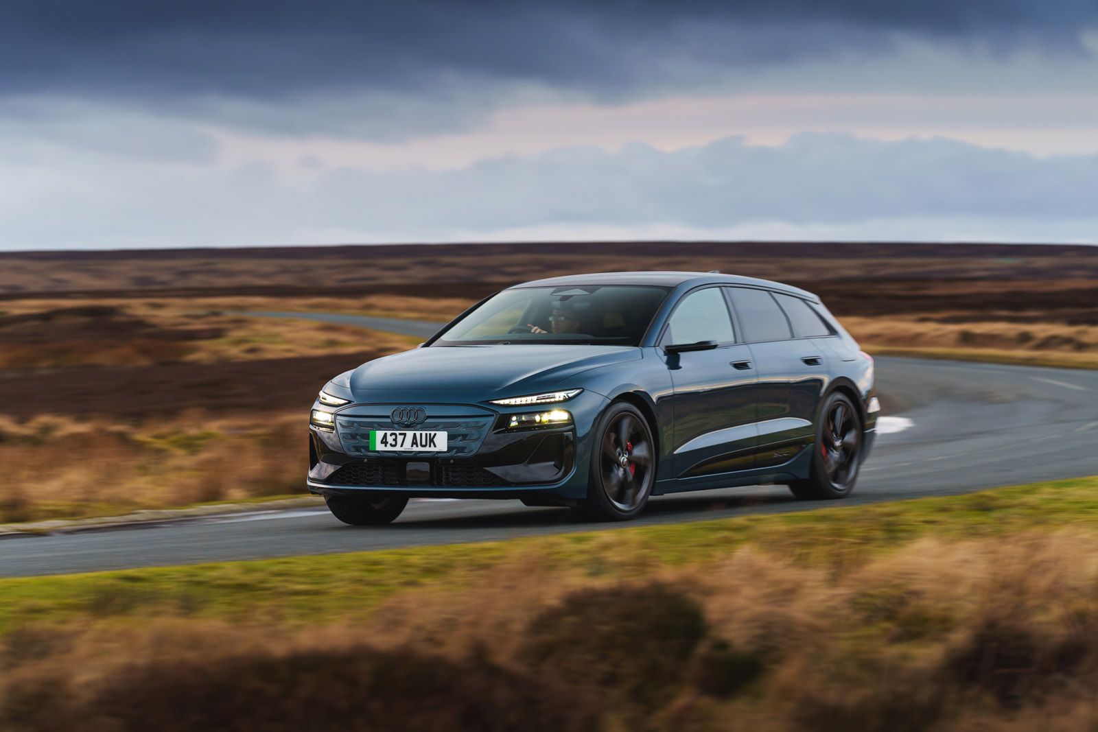 A sleek dark gray car drives on a winding road through a barren landscape under a cloudy sky.