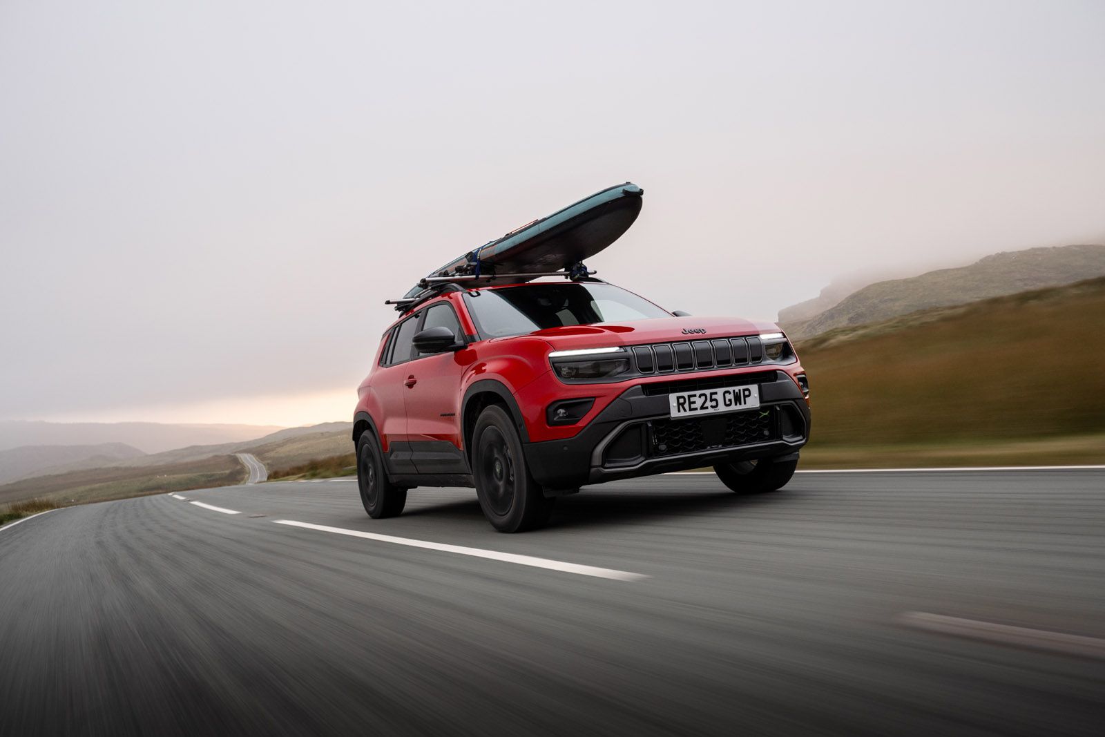 Red SUV with a roof rack carrying a kayak, driving on an empty road through a misty landscape, under a cloudy sky.