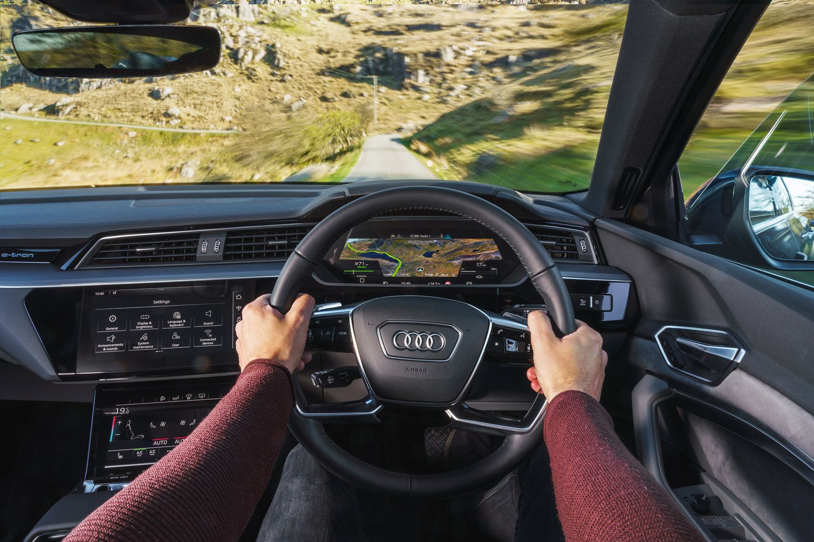 Driver's view inside a modern car, showing hands on the steering wheel, digital dashboard, and countryside road ahead.