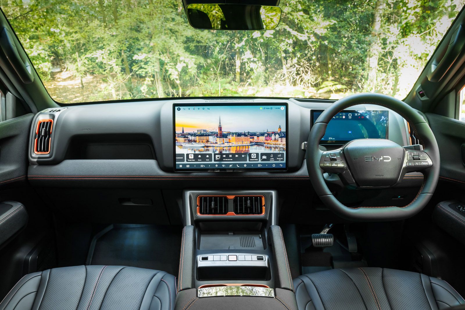 Car interior with steering wheel, two digital displays, leather seats, and a scenic screen image. Greenery visible through the windshield.