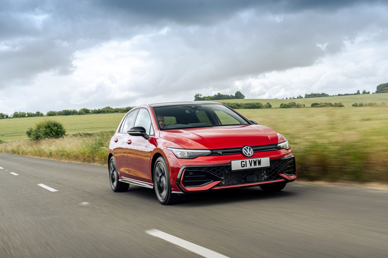 A red Volkswagen car driving on a countryside road with green fields and cloudy skies in the background.