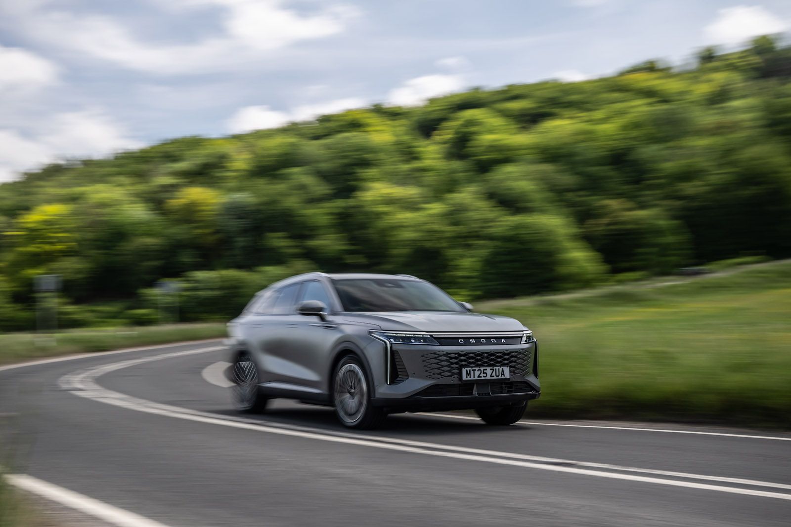 Sleek silver car speeding on a curvy road, surrounded by lush green trees under a partly cloudy sky.