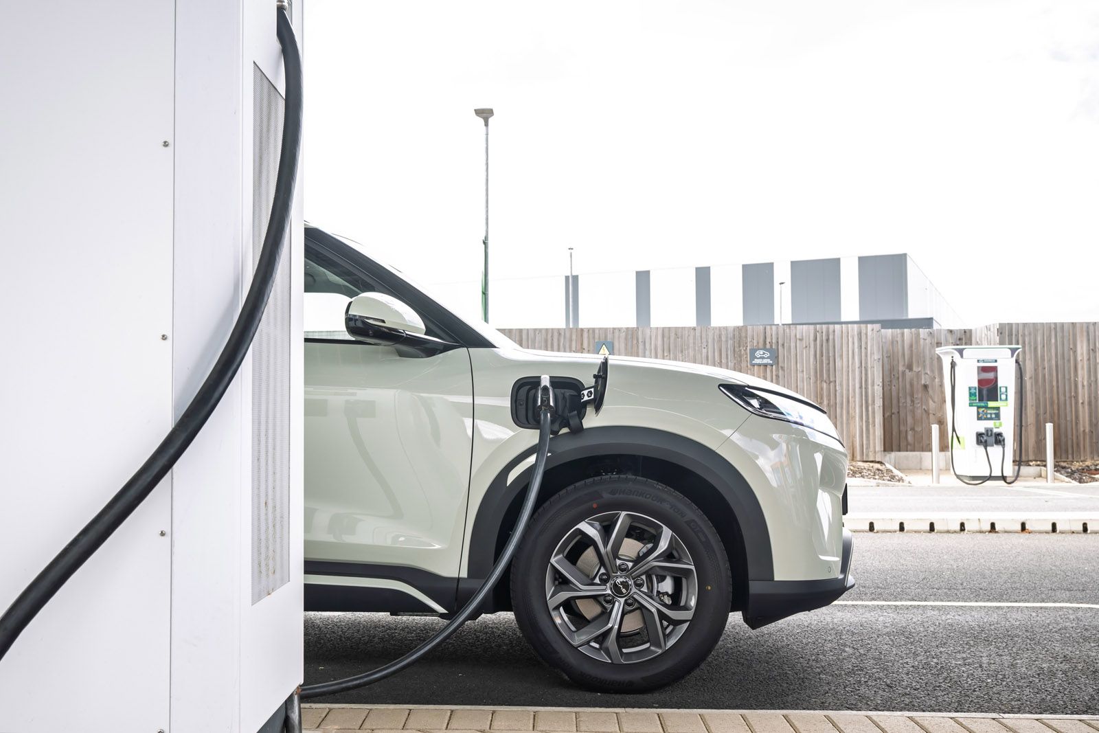 Electric SUV charging at a station, plugged into a charger on a cloudy day, with another charging point visible in the background.