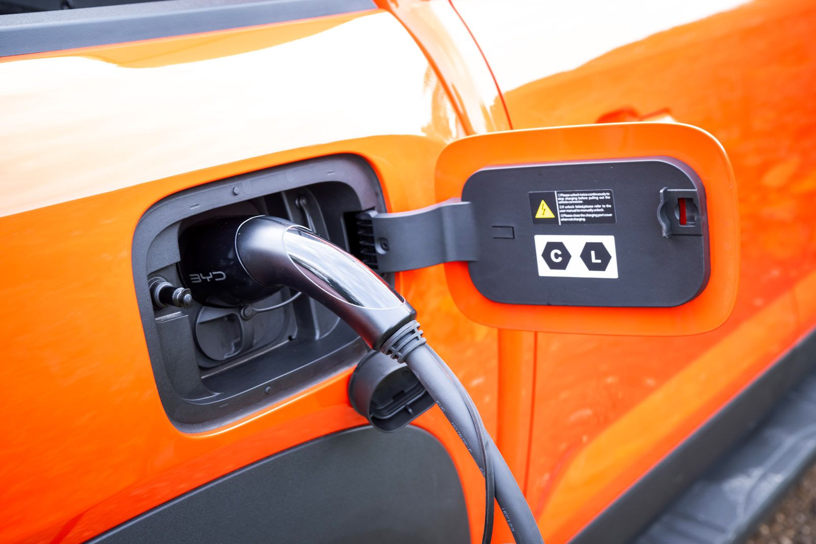 An orange electric vehicle plugged in for charging, with a close-up of the charging port and cable.
